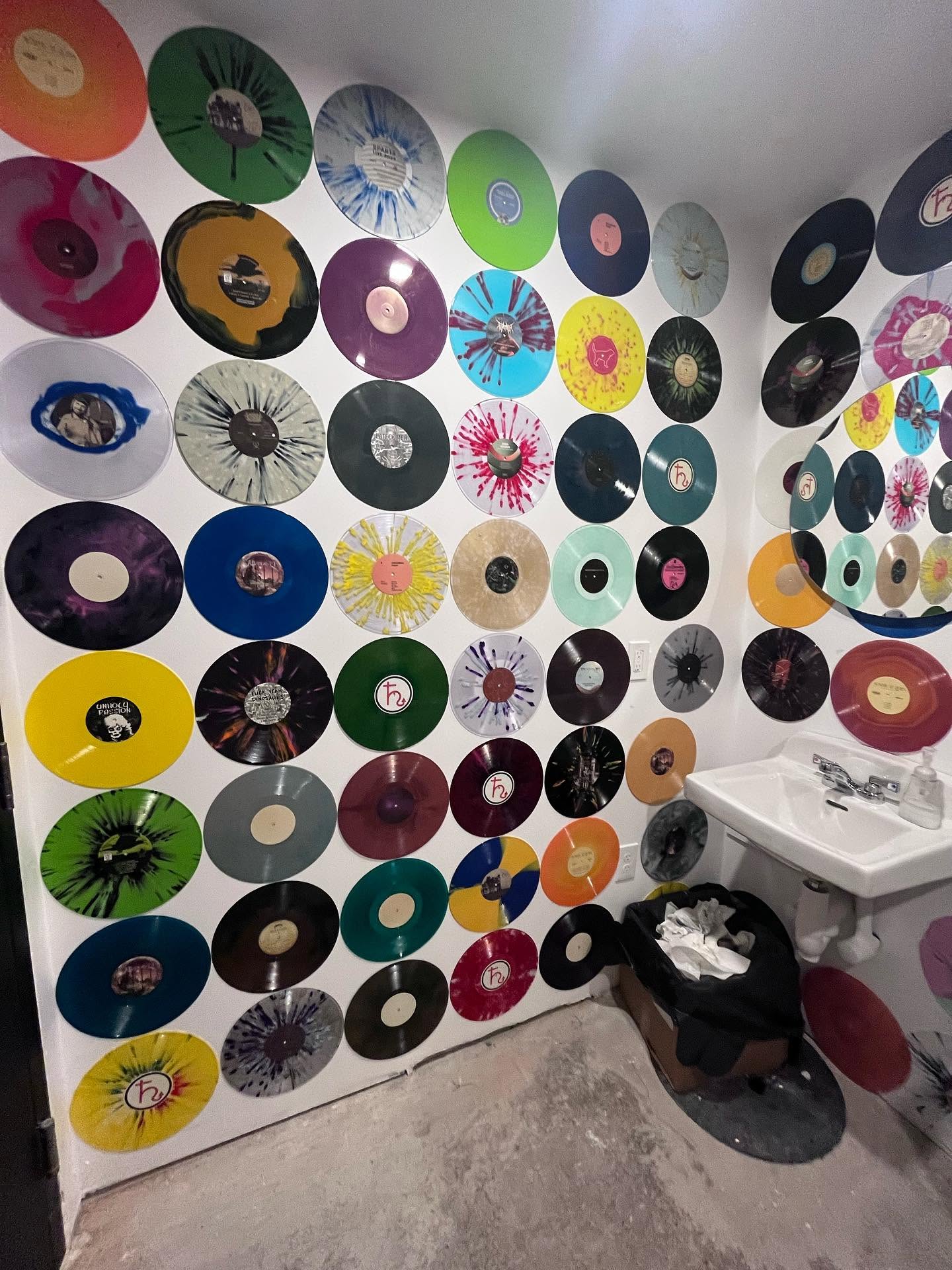 A wall covered with many colorful vinyl records mounted as decorative art in a bathroom or studio space, with a white sink and concrete floor visible.