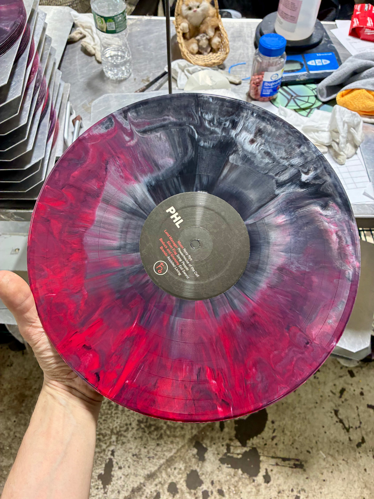 A hand holds a large vinyl record with a marbled red, pink, and black resin swirl pattern.