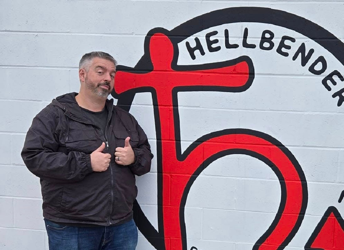 Man giving a thumbs-up in front of a wall with 'Hellbender' graffiti.